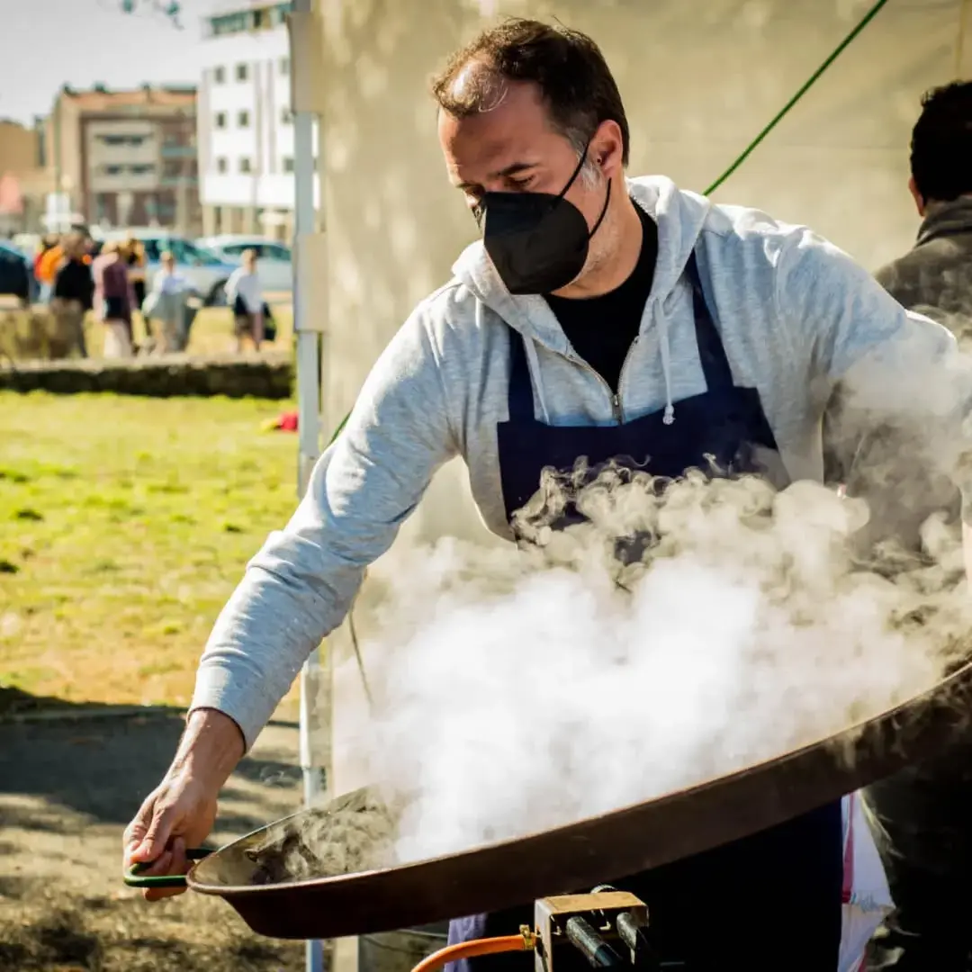 Chef cocinando una gran paella al aire libre en un evento gastronómico. El vapor se eleva mientras la paella se cocina a fuego vivo, creando una experiencia auténtica y deliciosa. Un momento perfecto para disfrutar de la cocina en directo Cuiner preparant una paella a l’aire lliure amb vapor sortint de la paella