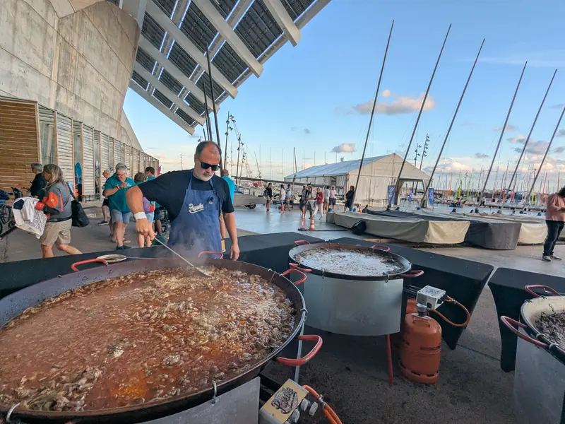 Cocinero sirviendo paellas gigantes en un puerto con veleros
